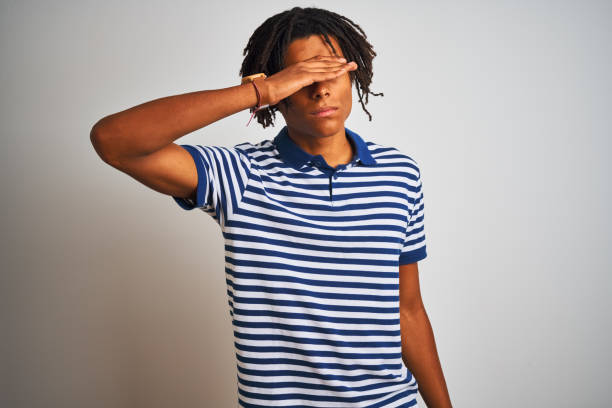 istock Afro man with dreadlocks wearing striped blue polo standing over isolated white background covering eyes with hand, looking serious and sad. Sightless, hiding and rejection concept 1162384264