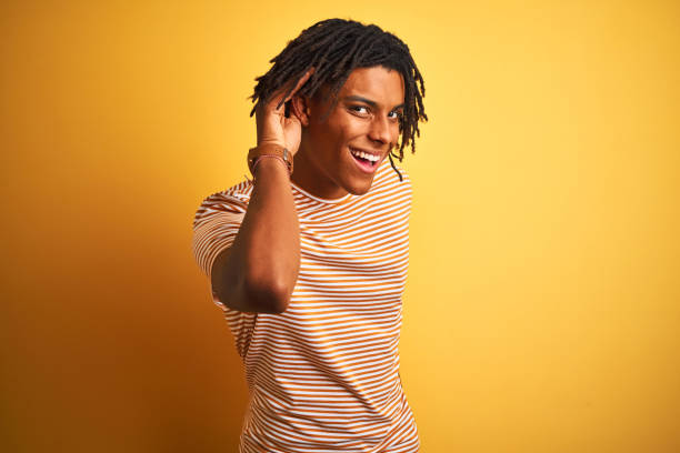 istock Afro man with dreadlocks wearing striped t-shirt standing over isolated yellow background smiling with hand over ear listening an hearing to rumor or gossip. Deafness concept. 1162384657