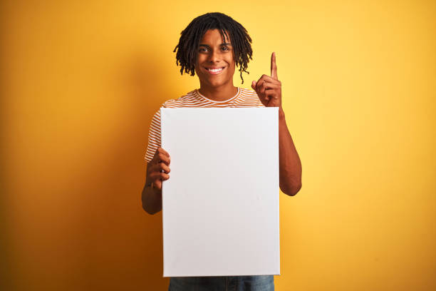 istock Afro american man with dreadlocks holding banner over isolated yellow background surprised with an idea or question pointing finger with happy face, number one 1162385682