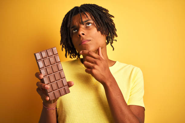 istock Afro american man with dreadlocks eating chocolate bar over isolated yellow background serious face thinking about question, very confused idea 1162386750
