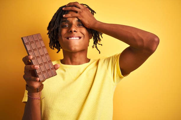 istock Afro american man with dreadlocks eating chocolate bar over isolated yellow background stressed with hand on head, shocked with shame and surprise face, angry and frustrated. Fear and upset for mistake. 1162386788