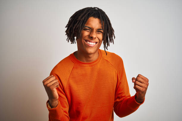 孤立した白い背景の上にオレンジ色のセーターを着たドレッドロックを持つアフロアメリカンの男は、腕を上げ、笑顔で成功のために叫んで勝者のジェスチャーをやって非常に幸せで興奮し� - men sweater excitement satisfaction ストックフォトと画像