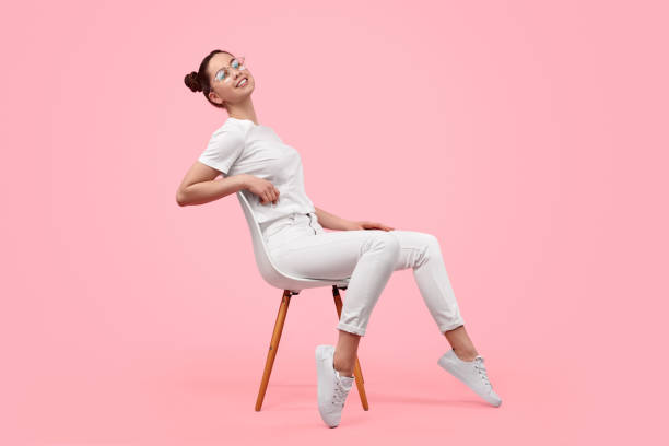 Relaxed teen girl sitting on chair Side view of cheerful female teenager in stylish white outfit smiling and leaning back while sitting on chair against pink background girl sitting stock pictures, royalty-free photos & images