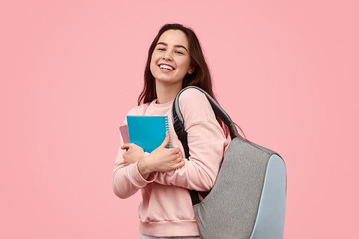 Carefree teen girl with backpack smiling and embracing notepads during studies against pink background