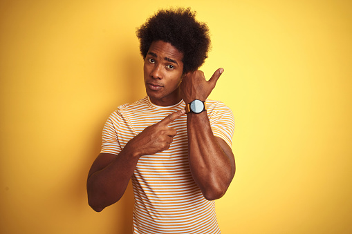 American man with afro hair wearing striped t-shirt standing over isolated yellow background In hurry pointing to watch time, impatience, looking at the camera with relaxed expression