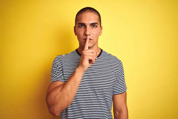 Photo of Young handsome man wearing striped t-shirt over yellow isolated background asking to be quiet with finger on lips. Silence and secret concept.