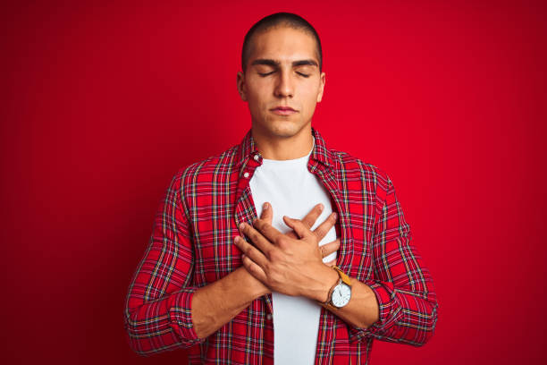 istock Young handsome man wearing a shirt using watch over red isolated background smiling with hands on chest with closed eyes and grateful gesture on face. Health concept. 1162405886