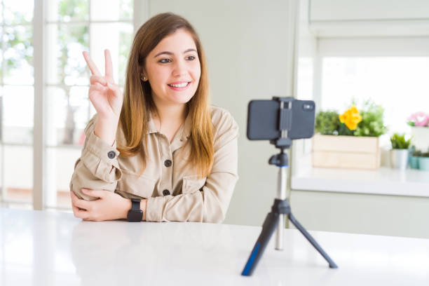 istock Beautiful young woman doing online video call using smartphone webcam smiling with happy face winking at the camera doing victory sign. Number two. 1162408284