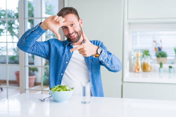 istock Handsome man eating fresh healthy salad smiling making frame with hands and fingers with happy face. Creativity and photography concept. 1162410567