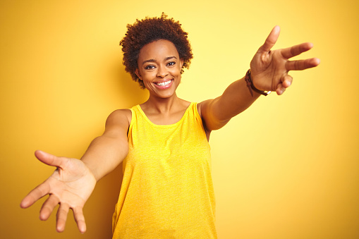Beauitul african american woman wearing summer t-shirt over isolated yellow background looking at the camera smiling with open arms for hug. Cheerful expression embracing happiness.