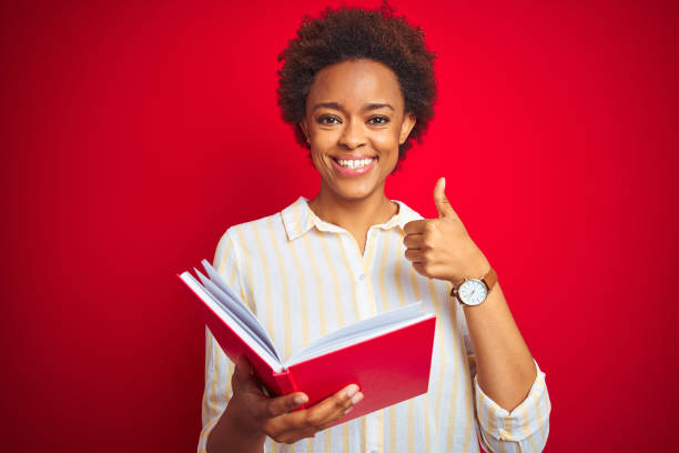 アフリカ系アメリカ人女性は、okサインをやって大きな笑顔で幸せな赤い孤立した背景の上に本を読んで、指で親指、優れた兆候 - thumbs up book smiling student ストックフォトと画像