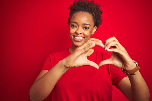 giovane bella donna afroamericana con i capelli afro su sfondo rosso isolato sorridente in amore facendo forma di simbolo del cuore con le mani. concetto romantico. - heart shape healthy lifestyle valentines day romance foto e immagini stock