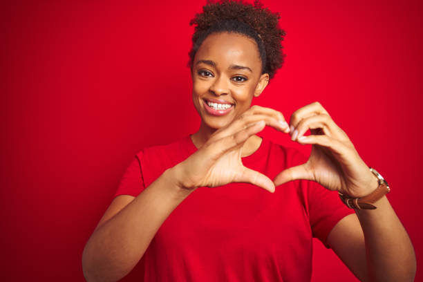 孤立した赤い背景の上にアフロヘアを持つ若い美しいアフリカ系アメリカ人女性は、手でハートシンボル形状をやって愛に微笑んでいます。ロマンチックなコンセプト。 - love red symbol valentines day ストックフォトと画像