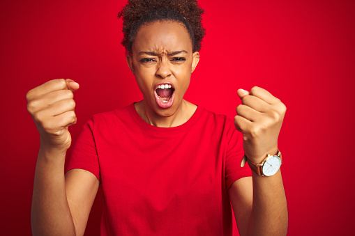 Young beautiful african american woman with afro hair over isolated red background angry and mad raising fists frustrated and furious while shouting with anger. Rage and aggressive concept.
