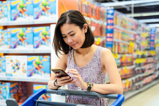 женщина проверки список покупок по мобильному телефону, потянув тележки для покупок в супермаркете - store on the phone supermarket sale стоковые фото и изображения