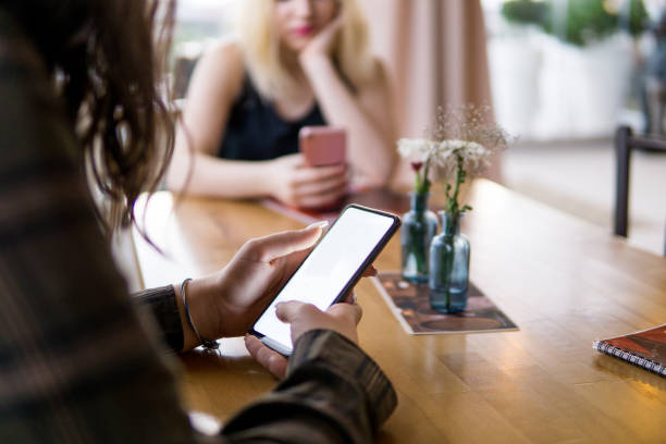 Girl using a mobile phone while out with her friend in a cafe, using a smart phone indoor to send a text message, checking social media, chatting online with wifi wireless
