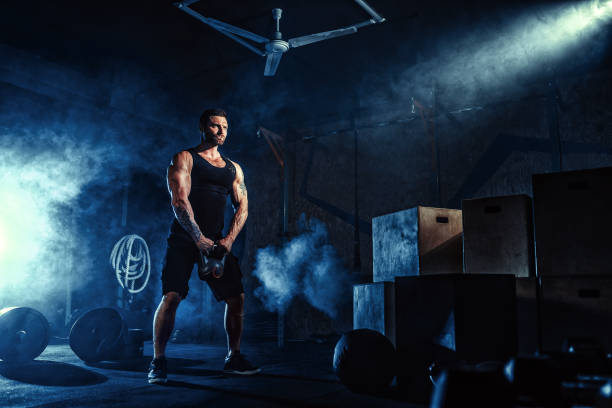 Muscular attractive caucasian bearded man lifting two kettlebells in a gym. Weight plates, dumbbell and tires in background Muscular attractive caucasian bearded tasttoed man lifting kettlebell in a gym. Weight plates, dumbbell and tires in smoke background. circuit training stock pictures, royalty-free photos & images