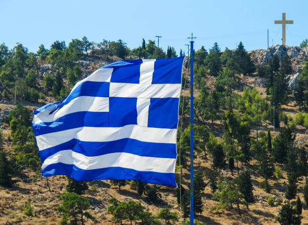 the national flag of greece on the flagpole develops in the wind against the background of a hill with green trees and a cross on top. greek island of kalymnos. - greece blue forest national landmark imagens e fotografias de stock