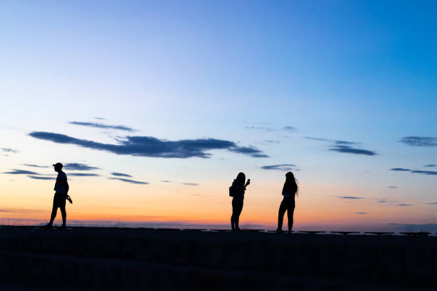 silhouettes de deux femmes qui prennent la photo sur le coucher du soleil et un homme seul - shadow focus on shadow women sunset photos et images de collection