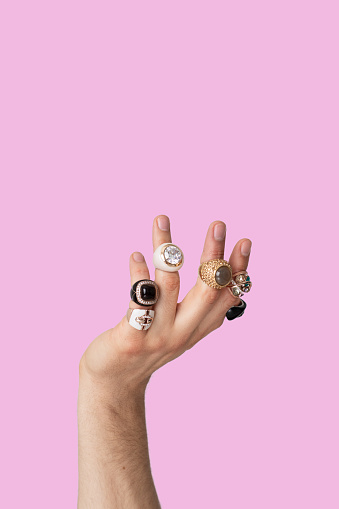 Close-up of man hand with female jewelry over pink background. Isolated man hand wearing woman rings on fingers, with copy space. Cross dressing unrecognizable person concept with negative space.