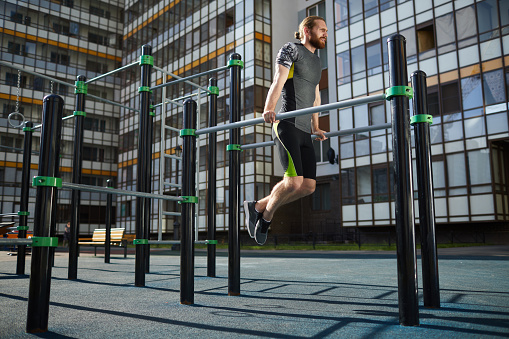 Tireless muscular athlete with red beard hanging on parallel bars while practicing pull-ups on training ground