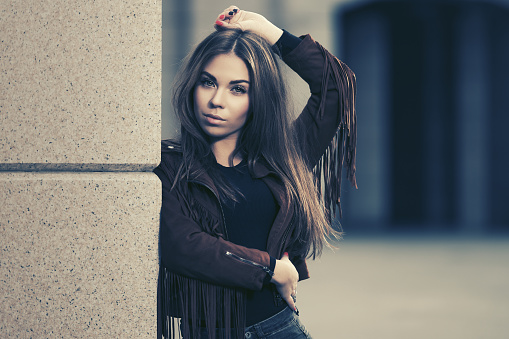 Young woman with long straight hairs leaning on the wall  Stylish fashion model wearing leather fringe suede jacket