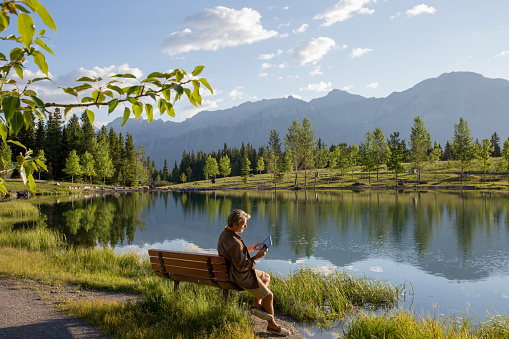 He is sitting on a park bench and surrounded by trees and mountains