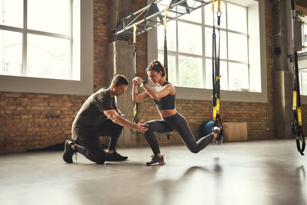 Doing squat exercise. Confident young personal trainer is showing slim athletic woman how to do squats with Trx fitness straps while training at gym. stock photo