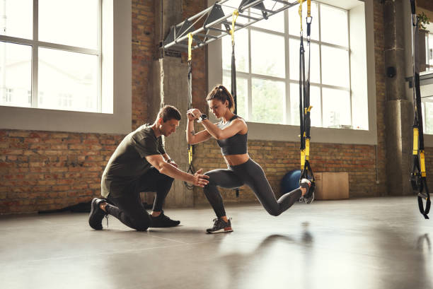 robi squat ćwiczenia. pewny siebie młody trener osobisty pokazuje szczupłą atletyczną kobietę, jak robić przysiady z paskami fitness trx podczas treningu na siłowni. - athlete zdjęcia i obrazy z banku zdjęć