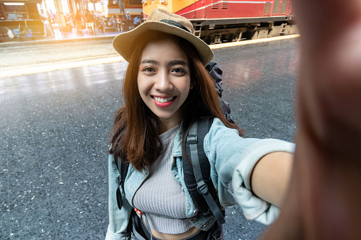 Attractive young Asian woman traveler with backpack taking a photo or selfie in train station. Travel lifestyle concept.
