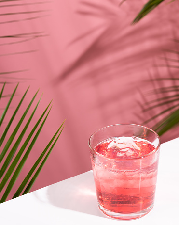 Pink Summer cold alcoholic cocktail on a white bar table. Green palm tree leaves on the background. Copy space for your text. Hard light