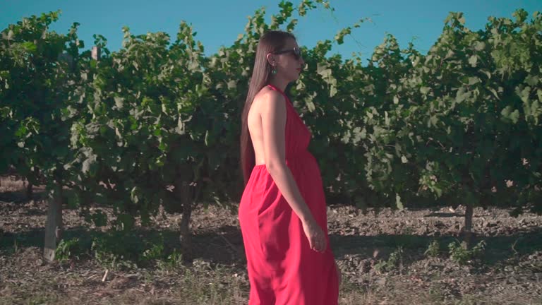 A pregnant girl in a red dress is walking through the vineyard. A pregnant girl with long hair in glasses.