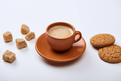 delicious coffee with foam in cup on saucer near brown sugar and cookies on white background