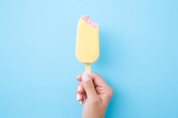 jeune main de femme retenant la crème glacée rose avec le glaçage blanc de chocolat sur le fond bleu pastel. de la nourriture mordue. closeup. vue du haut. - batonnet glace photos et images de collection