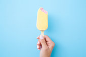 Young woman hand holding pink ice cream with white chocolate glaze on pastel blue background. Bitten food. Closeup. Top view.
