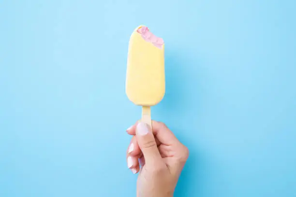 Photo of Young woman hand holding pink ice cream with white chocolate glaze on pastel blue background. Bitten food. Closeup. Top view.
