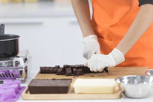 Beautiful Asian female cook in orange apron using knife to cut chocolate bar into small pieces to make many shape chocolate in modern kitchen.