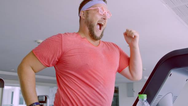 divertente maschio grasso con occhiali rosa e in una maglietta rosa è impegnato su un tapis roulant in palestra raffigurante una ragazza. 4k - irony foto e immagini stock
