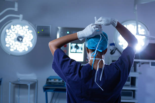 Female surgeon wearing surgical mask on in operating room at hospital Rear view of young African-american female surgeon putting her surgical mask on in operating room at hospital. Shot in real medical hospital with doctors nurses and surgeons in authentic setting operating room photos stock pictures, royalty-free photos & images