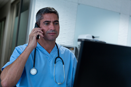 Front view of Caucasian male surgeon talking on mobile phone while working on computer at desk in hospital. Shot in real medical hospital with doctors nurses and surgeons in authentic setting