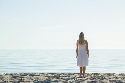 Young blonde woman in white dress standing alone on sand and staring at calm sea and light blue sky. Fresh air. Empty place for emotional, sentimental, inspirational text, quote or sayings. Back view.