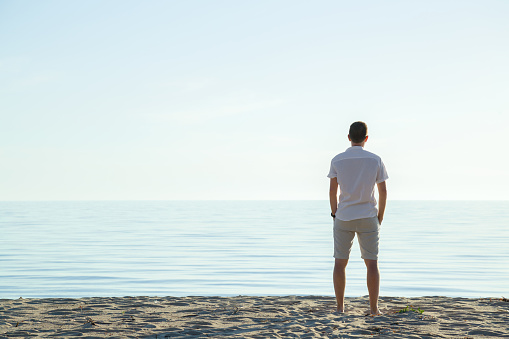 Young man in white clothes standing alone on sand and staring at calm sea and light blue sky. Fresh air. Empty place for emotional, sentimental, inspirational text, quote or sayings. Back view.