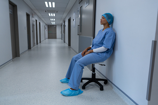 Side view of tensed Mixed race female surgeon eyes closed sitting on stool in the corridor at hospital. Shot in real medical hospital with doctors nurses and surgeons in authentic setting