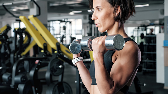 Portrait of athletic mature woman is making set of reps exercise for biceps with dumbbells in hands in gym. She is lifting dumbbells. Training and sport concept.