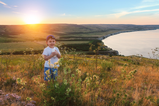 Smiling little child is holding a glass of milk at the picturesque nature background. Funny baby. Amazing summertime.