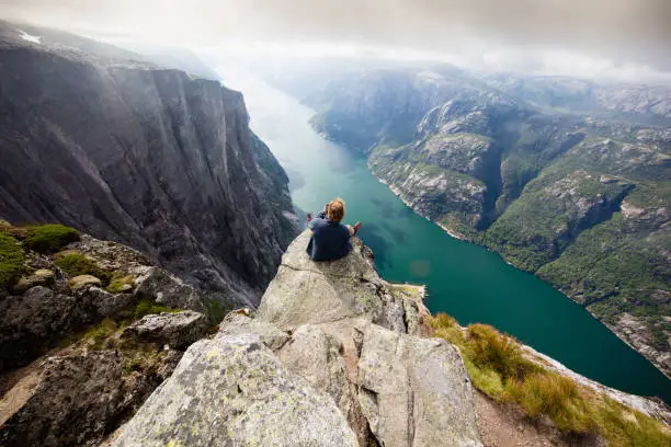 Hiker sitting on Kjeragnasen over Lyseford in Norway taking pictures with smartphone