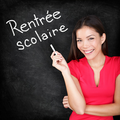 Rentree Scolaire - French teacher Back to School written in French on blackboard by woman teacher holding chalk. Smiling happy woman teaching French language or university student back in college.