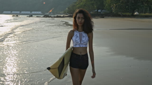 Asian teenage girl holding a surfboard on the beach with beautiful sea in Thailand.