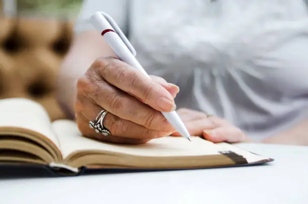 Photo of Senior woman writing with pencil on open note book.