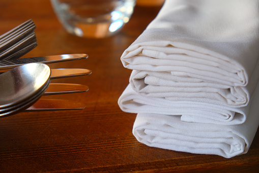 A restaurant table on the left are knives, forks and spoons stacked ontop of each other on the right four white linen napkins, wooden table, warm colours.